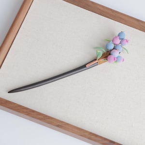 May include: A dark wooden hair stick with a decorative cluster of crocheted berries in shades of pink and blue, accented with green leaves. The hair accessory is set against a neutral background within a wooden frame.