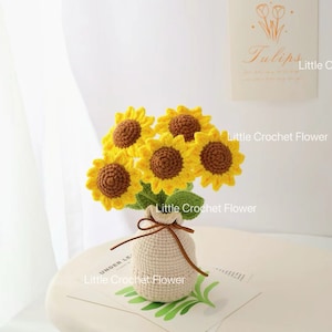 May include: A crocheted bouquet of sunflowers in a small beige bag tied with a brown string. The sunflowers have yellow petals and brown centres. The bag sits on a white surface with a small card that says "Tulips".