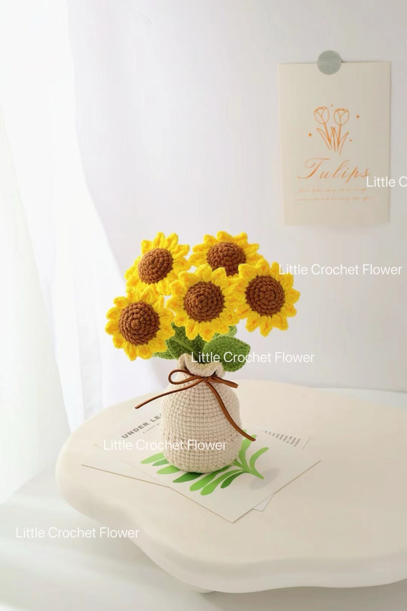May include: A crocheted bouquet of sunflowers in a small beige bag tied with a brown string. The sunflowers have yellow petals and brown centres. The bag sits on a white surface with a small card that says "Tulips".