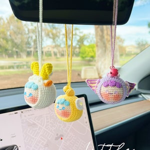 Puede incluir: Tres juguetes de peluche tejidos a crochet colgando del espejo retrovisor de un coche. Los juguetes son un cordero amarillo y blanco, un pollito amarillo y un unicornio morado y blanco. Todos los juguetes sonr&iacute;en y tienen ojos grandes. La palabra "little" est&aacute; escrita en blanco en la parte inferior de la imagen.