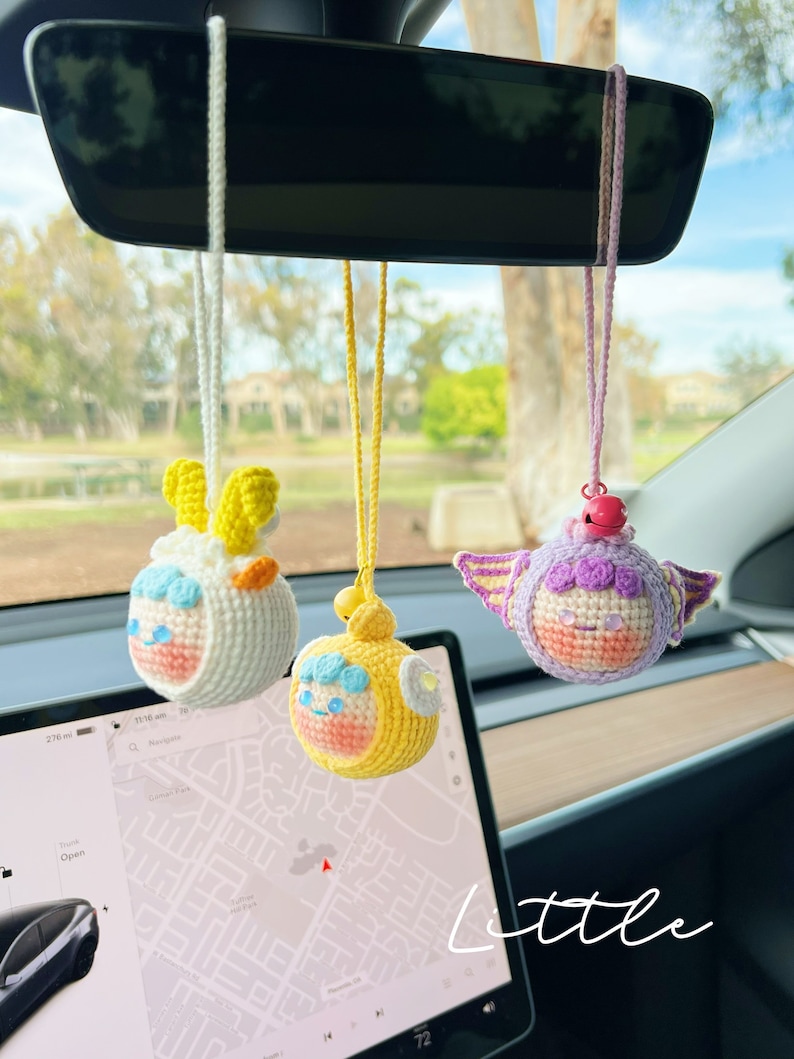 Puede incluir: Tres juguetes de peluche tejidos a crochet colgando del espejo retrovisor de un coche. Los juguetes son un cordero amarillo y blanco, un pollito amarillo y un unicornio morado y blanco. Todos los juguetes sonr&iacute;en y tienen ojos grandes. La palabra "little" est&aacute; escrita en blanco en la parte inferior de la imagen.