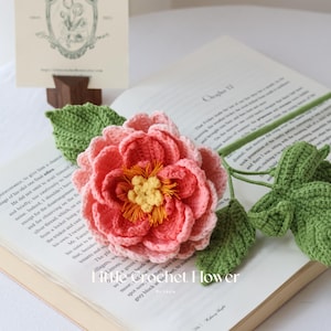 K&ouml;nnte beinhalten: Eine handgefertigte H&auml;kelblume in Rosa- und Korallt&ouml;nen, mit gelber Mitte und gr&uuml;nen Bl&auml;ttern. Die Blume liegt auf einem offenen Buch. Der Text "Crochet Flower" ist sichtbar.