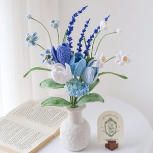 May include: A crochet flower bouquet featuring blue and white tulips, small light blue blossoms, and tall dark blue spikes, all with green stems and leaves, arranged in a white textured vase with a small card displaying a floral illustration.