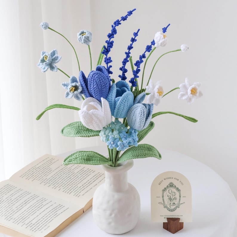 May include: A crochet flower bouquet featuring blue and white tulips, small light blue blossoms, and tall dark blue spikes, all with green stems and leaves, arranged in a white textured vase with a small card displaying a floral illustration.