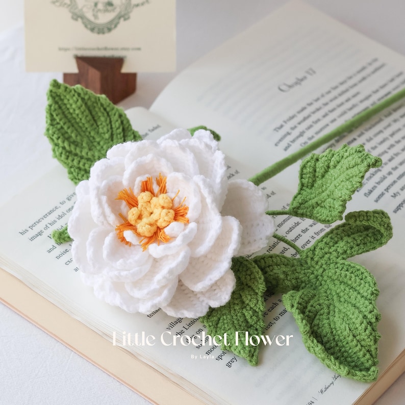 K&ouml;nnte beinhalten: Eine handgefertigte H&auml;kelblume mit wei&szlig;en Bl&uuml;tenbl&auml;ttern, einem orangefarbenen und gelben Zentrum und gr&uuml;nen Bl&auml;ttern. Die Blume liegt auf einem offenen Buch. Der Text "Little Crochet Flower" ist sichtbar.