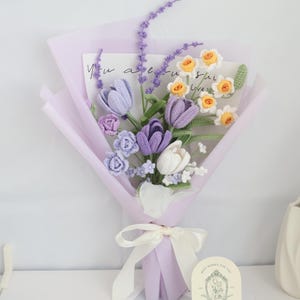 May include: A crocheted bouquet of purple and white tulips, yellow daffodils, and lavender sprigs. The bouquet is wrapped in light purple tissue paper and tied with a white satin ribbon. A small card with the text "Best Wishes for You" is in the foreground.