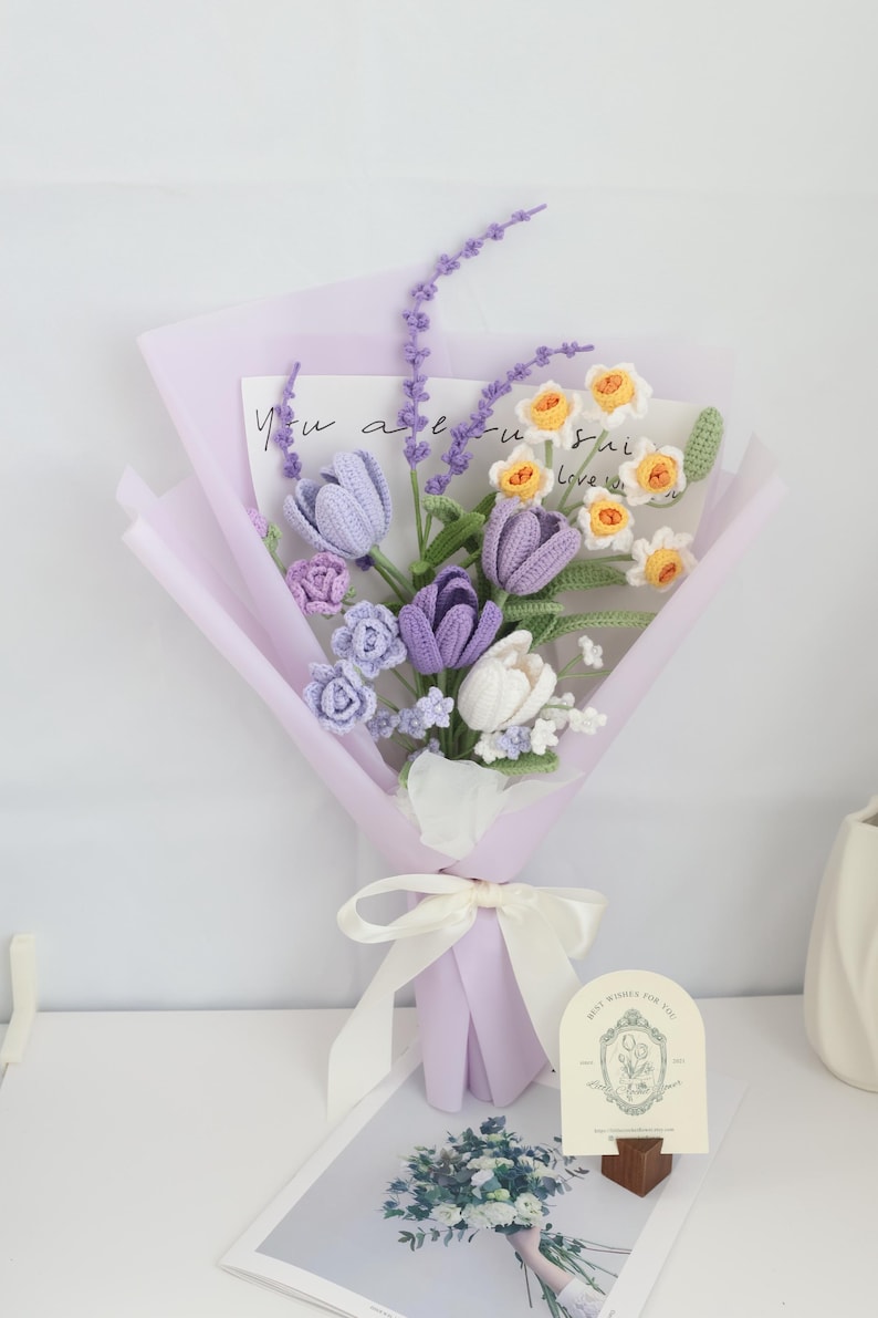 May include: A crocheted bouquet of purple and white tulips, yellow daffodils, and lavender sprigs. The bouquet is wrapped in light purple tissue paper and tied with a white satin ribbon. A small card with the text "Best Wishes for You" is in the foreground.