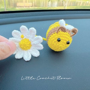May include: A crocheted yellow and brown bee with black eyes and a white crocheted flower. The bee and flower are on a dark gray surface. The text "Little Crochet Flower" is written below the bee and flower.