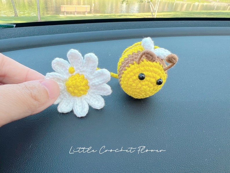 May include: A crocheted yellow and brown bee with black eyes and a white crocheted flower. The bee and flower are on a dark gray surface. The text "Little Crochet Flower" is written below the bee and flower.