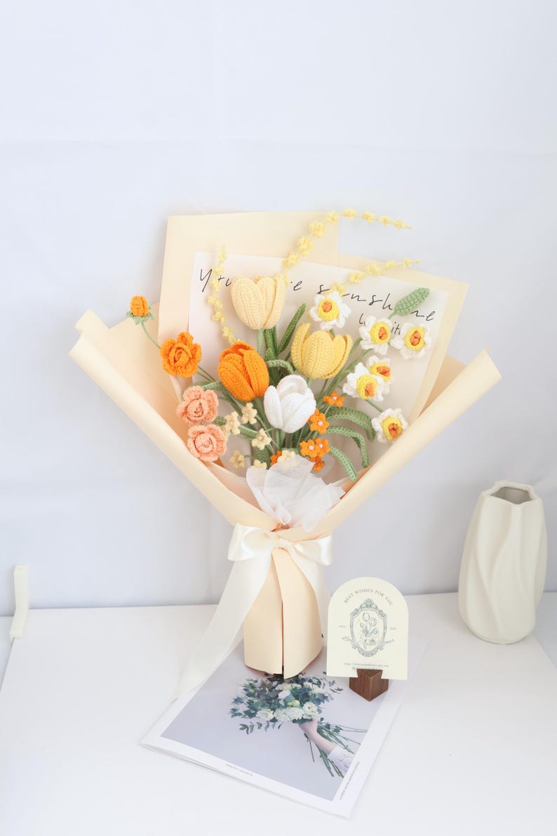 May include: A crocheted bouquet of yellow, orange, and white flowers in a cream paper wrapping. The bouquet is tied with a white ribbon and has a small card that says "You're sunshine with it".