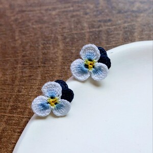 May include: A pair of crocheted flower earrings with white petals and blue and yellow centers. The earrings are on a white surface.