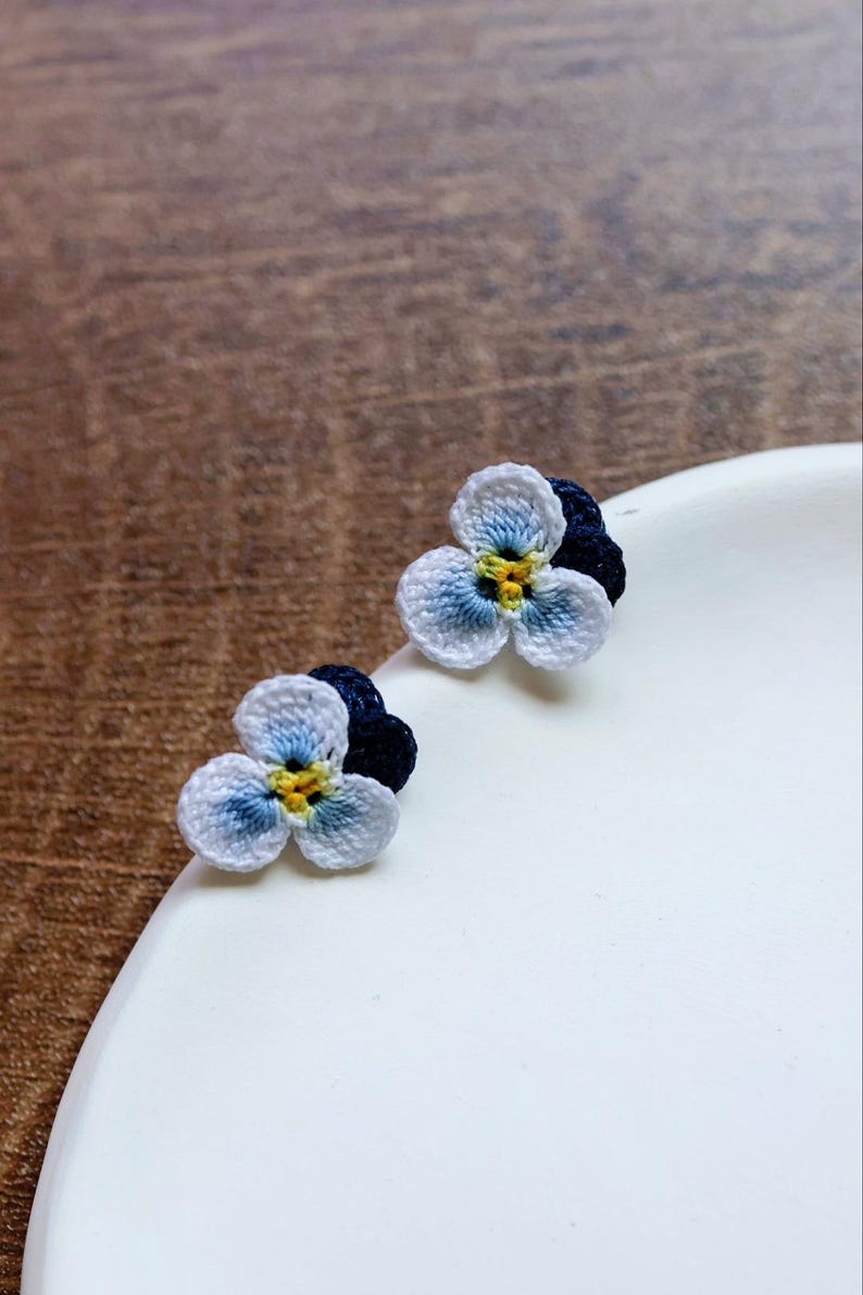 May include: A pair of crocheted flower earrings with white petals and blue and yellow centers. The earrings are on a white surface.