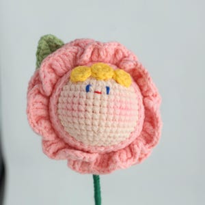 Puede incluir: Una flor de ganchillo hecha a mano con una cara sonriente. La flor tiene p&eacute;talos rosas, una cara rosa claro, cabello amarillo y ojos azules. Tambi&eacute;n se ven un tallo y una hoja verdes. Ideal para la decoraci&oacute;n del hogar.