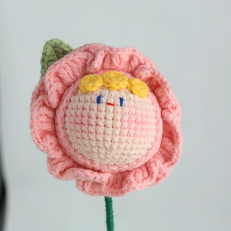 Puede incluir: Una flor de ganchillo hecha a mano con una cara sonriente. La flor tiene p&eacute;talos rosas, una cara rosa claro, cabello amarillo y ojos azules. Tambi&eacute;n se ven un tallo y una hoja verdes. Ideal para la decoraci&oacute;n del hogar.
