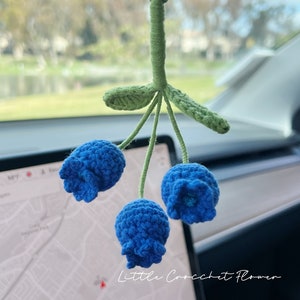 K&ouml;nnte beinhalten: Eine blaue geh&auml;kelte Blume mit gr&uuml;nen Bl&auml;ttern, die an einem R&uuml;ckspiegel h&auml;ngt. Die Blume hat drei Bl&uuml;ten und der Text "Little Crochet Flower" ist auf dem Bild zu sehen.