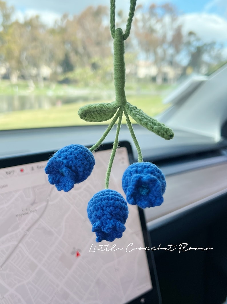 K&ouml;nnte beinhalten: Eine blaue geh&auml;kelte Blume mit gr&uuml;nen Bl&auml;ttern, die an einem R&uuml;ckspiegel h&auml;ngt. Die Blume hat drei Bl&uuml;ten und der Text "Little Crochet Flower" ist auf dem Bild zu sehen.