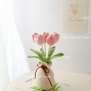 May include: A handmade crochet flower arrangement featuring pink and light pink tulips in a beige crocheted vase tied with a brown ribbon. The arrangement sits on a white table with a decorative card that says "Tulips".