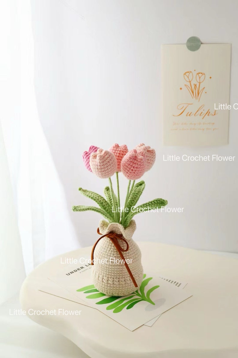 May include: A handmade crochet flower arrangement featuring pink and light pink tulips in a beige crocheted vase tied with a brown ribbon. The arrangement sits on a white table with a decorative card that says "Tulips".