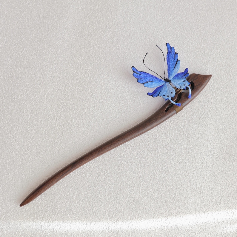 May include: A dark brown wooden hair stick with a decorative blue butterfly. The butterfly has detailed wings with varying shades of blue. The hair accessory is designed for elegant hairstyles.