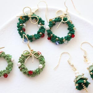 May include: Four pairs of handmade Christmas-themed earrings. Two pairs feature green crocheted wreaths with red and white beads. One pair features a green crocheted Christmas tree with white beads. The other pair features a green crocheted Christmas tree with white beads.