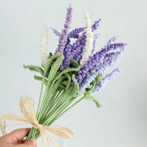 Pode incluir: Um buquê de flores de lavanda de crochê em tons de roxo, lavanda e branco, com folhas e caules verdes. Os caules são amarrados com uma fita de ráfia bege. Um item decorativo charmoso.