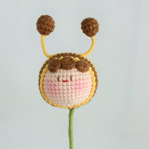 Puede incluir: Una abeja de ganchillo hecha a mano con un cuerpo redondo, mejillas rosadas y una peque&ntilde;a boca roja. La abeja tiene antenas marrones con puntas de pomp&oacute;n marrones y un cuerpo a rayas amarillas y marrones. La abeja est&aacute; unida a un tallo verde.