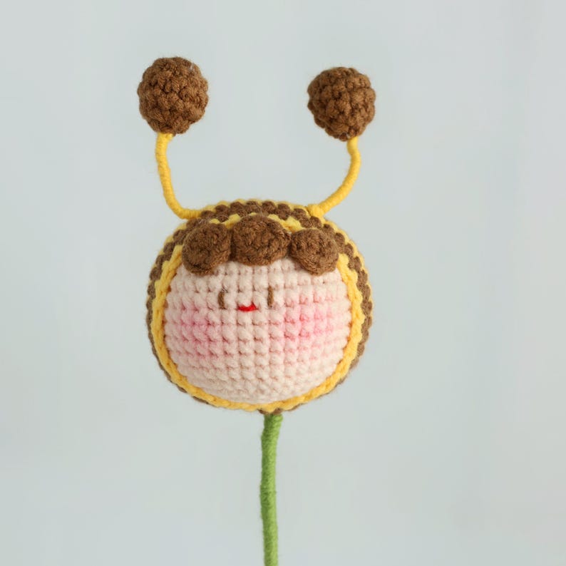 Puede incluir: Una abeja de ganchillo hecha a mano con un cuerpo redondo, mejillas rosadas y una peque&ntilde;a boca roja. La abeja tiene antenas marrones con puntas de pomp&oacute;n marrones y un cuerpo a rayas amarillas y marrones. La abeja est&aacute; unida a un tallo verde.