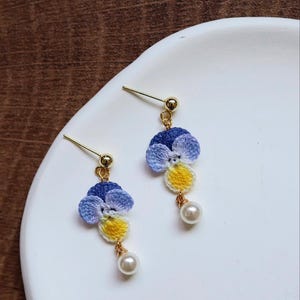 May include: A pair of dangle earrings featuring crocheted purple, blue, and yellow flowers with a pearl accent.