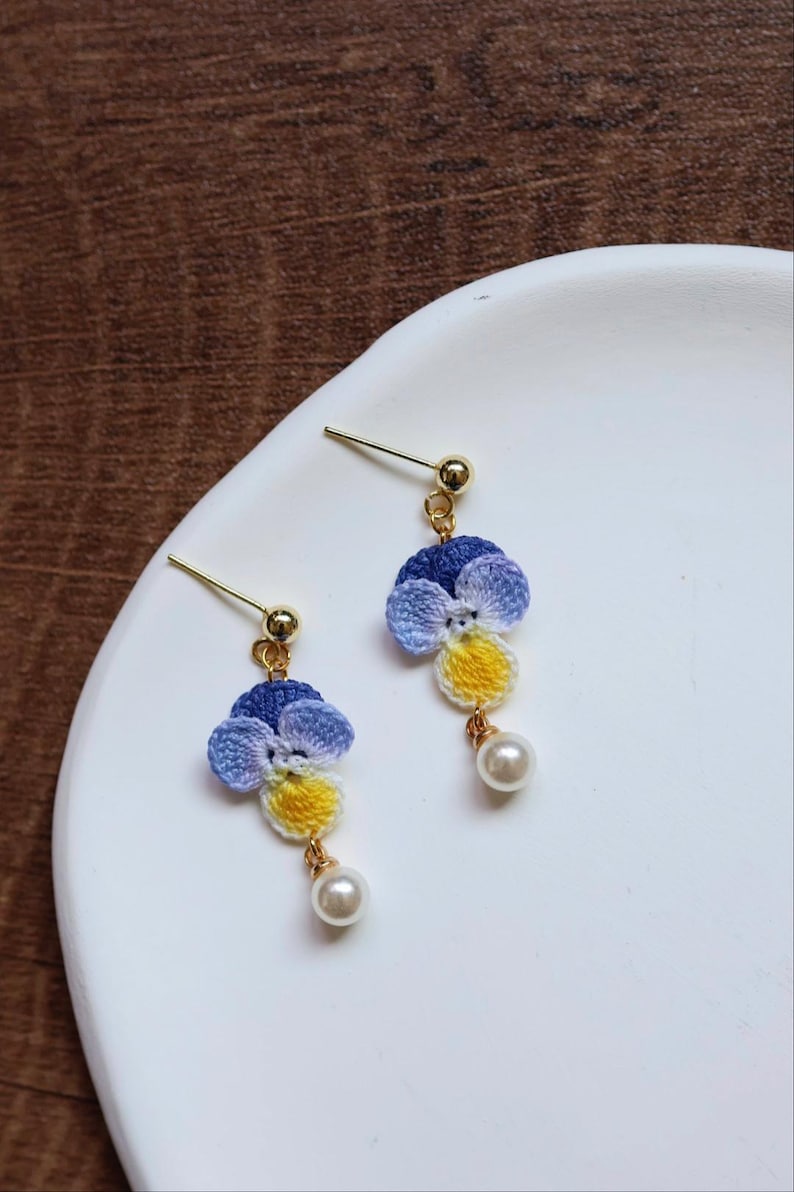 May include: A pair of dangle earrings featuring crocheted purple, blue, and yellow flowers with a pearl accent.