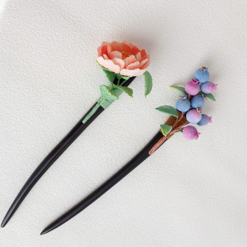 May include: Two decorative hair sticks. One features a peach-colored flower with green leaves, the other has a cluster of blue and pink berries with green leaves. Both have long, dark wooden stems.