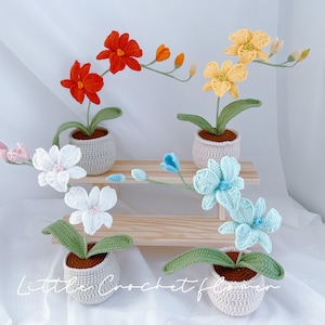 May include: Four crocheted flower arrangements in pots. The flowers are in shades of orange, yellow, white, and blue. The pots are a light gray color. The flowers are arranged on a wooden shelf.