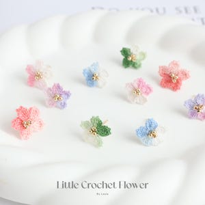 Op de afbeelding: Een verzameling handgemaakte gehaakte bloemoorbellen in verschillende kleuren, waaronder roze, blauw, groen en paars. Elke oorbel heeft een delicaat bloemmotief met goudkleurige kraalaccenten. De oorbellen worden gepresenteerd op een wit, golvend oppervlak. De tekst op de afbeelding luidt "Little Crochet Flower".