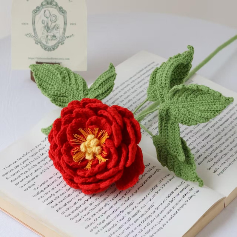 K&ouml;nnte beinhalten: Eine handgefertigte, rote H&auml;kelblume mit gelben Mittelteilen und gr&uuml;nen Bl&auml;ttern. Die Blume befindet sich auf einem langen gr&uuml;nen Stiel. Die Blume liegt auf einem offenen Buch.
