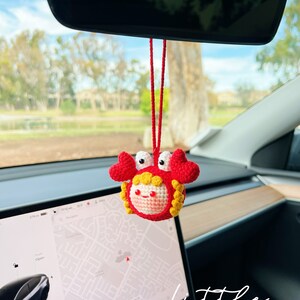 Puede incluir: Un cangrejo de crochet rojo y amarillo colgando del espejo retrovisor de un coche. El cangrejo tiene una cara sonriente y sostiene una cuerda roja. El texto "little" es visible en la esquina inferior derecha de la imagen.