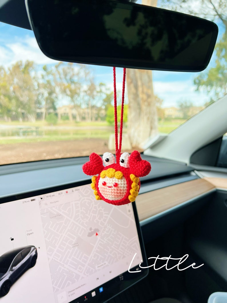 Puede incluir: Un cangrejo de crochet rojo y amarillo colgando del espejo retrovisor de un coche. El cangrejo tiene una cara sonriente y sostiene una cuerda roja. El texto "little" es visible en la esquina inferior derecha de la imagen.