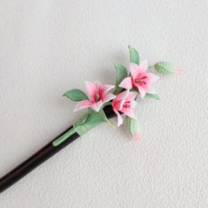May include: A handcrafted hair stick featuring a crochet design of pink lily flowers with green leaves and buds. The flowers have white accents and red centers. The hair stick is dark brown and wrapped with green thread.