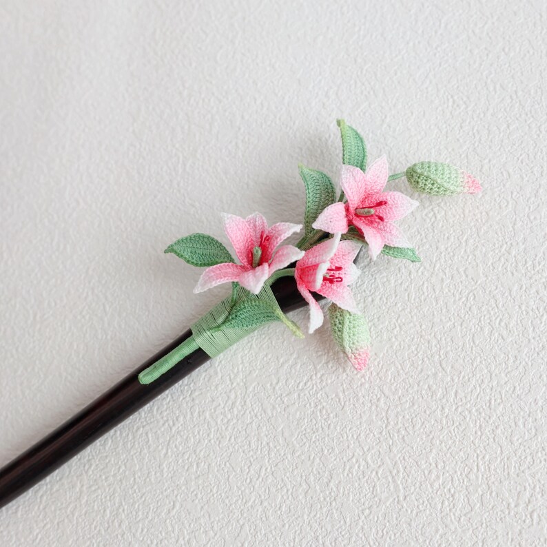 May include: A handcrafted hair stick featuring a crochet design of pink lily flowers with green leaves and buds. The flowers have white accents and red centers. The hair stick is dark brown and wrapped with green thread.