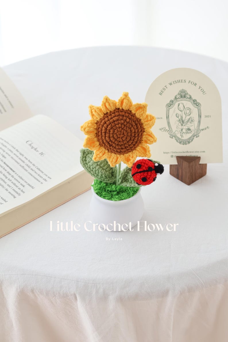 May include: A handmade crochet sunflower with a ladybug accent in a white pot. The sunflower has yellow petals and a brown center. The ladybug is red with black spots. The text "Little Crochet Flower" is at the bottom.