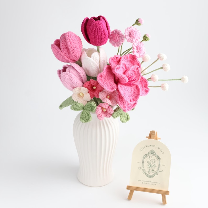 May include: A bouquet of crocheted flowers in shades of pink, magenta, and white, including tulips and small blossoms with green leaves, arranged in a white ribbed vase. Next to the vase is a small wooden easel holding a card with floral graphics.
