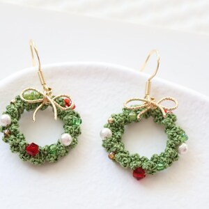 May include: A pair of green crocheted Christmas wreath earrings with red and white beads and a gold bow.