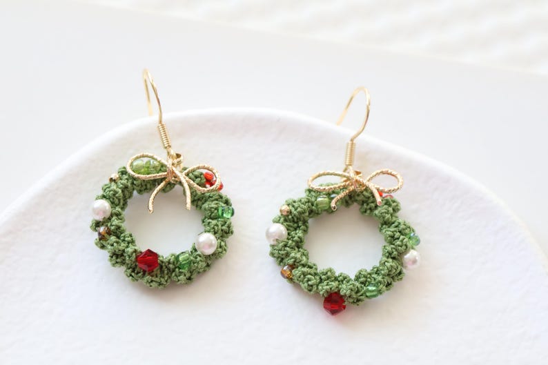 May include: A pair of green crocheted Christmas wreath earrings with red and white beads and a gold bow.