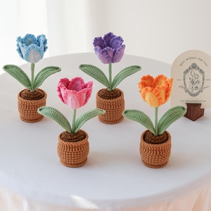 Può includere: Fiori di tulipano all'uncinetto nei toni del blu, rosa, viola e arancione, ognuno con foglie verdi e un vaso marrone. I fiori fatti a mano sono disposti su una superficie bianca. Un piccolo cartello recita "Best Wishes for You".