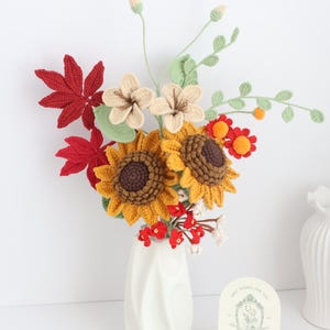 May include: A handcrafted crochet bouquet in a white vase. The arrangement features sunflowers, red maple leaves, and other colorful flowers. A small card with text is in front of the vase. The bouquet is a decorative item.