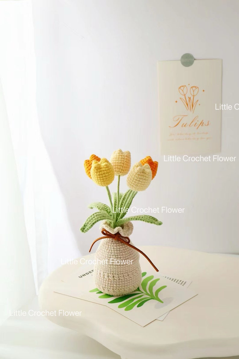 May include: A crocheted bouquet of tulips in a small vase. The flowers are yellow and orange, with green stems and leaves. The vase is beige and tied with a brown ribbon. A card with the word "Tulips" is in the background.