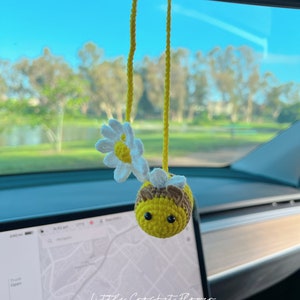 May include: A crocheted bee with a yellow body, brown head, and black eyes hangs from a yellow string in a car. The bee is holding a white crocheted daisy with a yellow center. The text "Little Crochet Flower" is visible in the image.