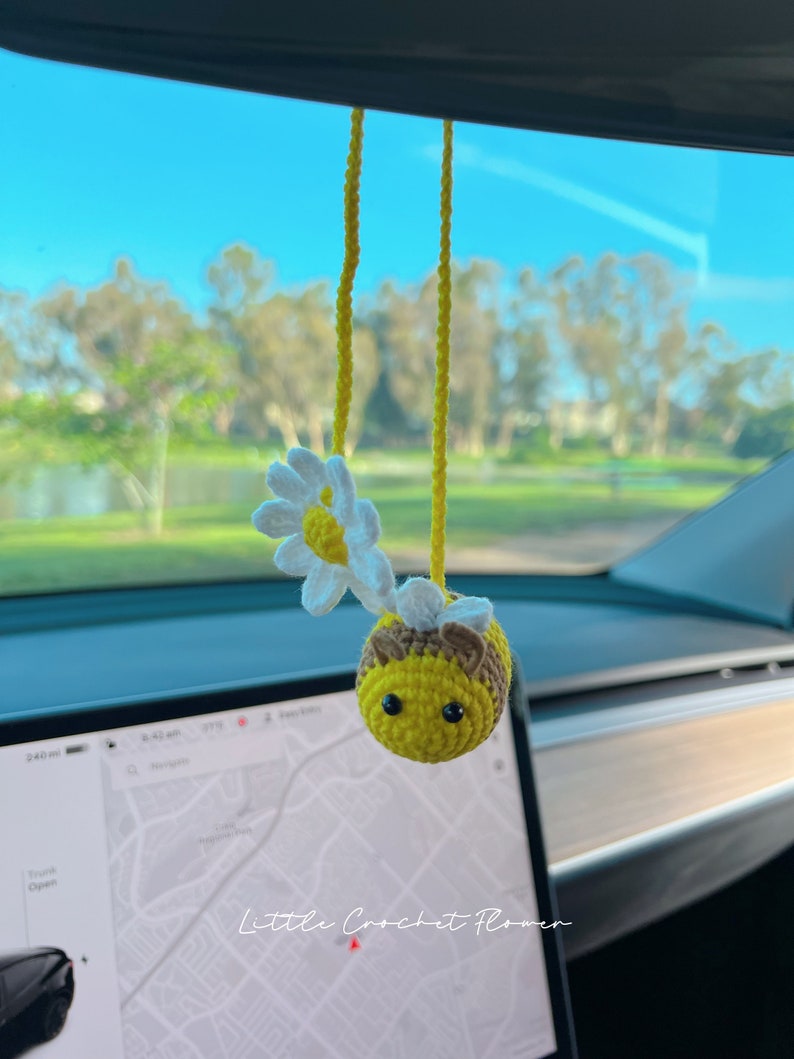 May include: A crocheted bee with a yellow body, brown head, and black eyes hangs from a yellow string in a car. The bee is holding a white crocheted daisy with a yellow center. The text "Little Crochet Flower" is visible in the image.