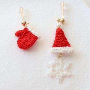 May include: A pair of Christmas-themed earrings. One earring features a red crocheted mitten, while the other earring features a red crocheted Santa hat with a white trim and a white crocheted snowflake dangling below.