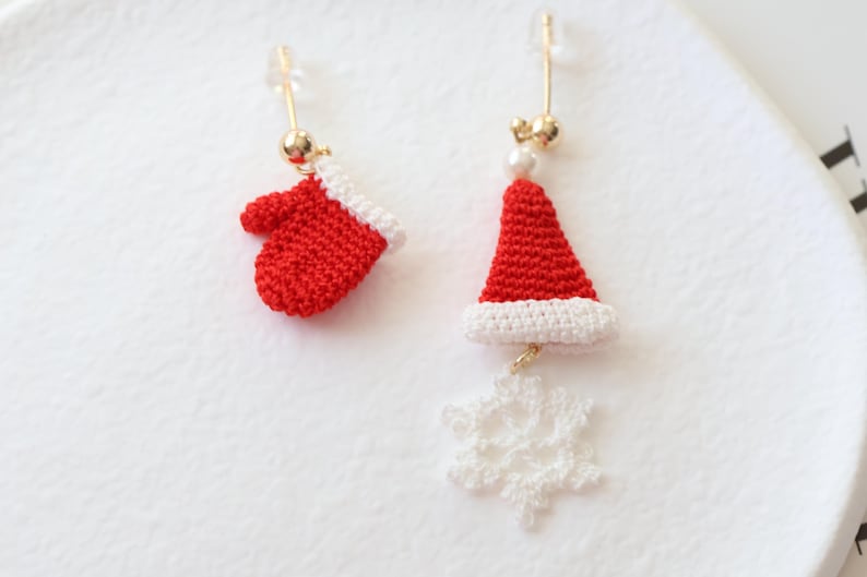 May include: A pair of Christmas-themed earrings. One earring features a red crocheted mitten, while the other earring features a red crocheted Santa hat with a white trim and a white crocheted snowflake dangling below.