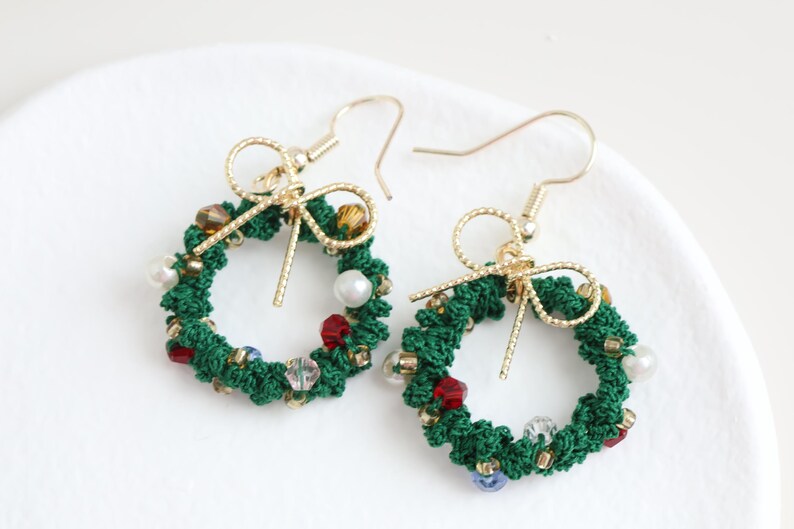 May include: A pair of green Christmas wreath earrings with gold hooks and a gold bow. The wreaths are decorated with red, white, and blue beads.