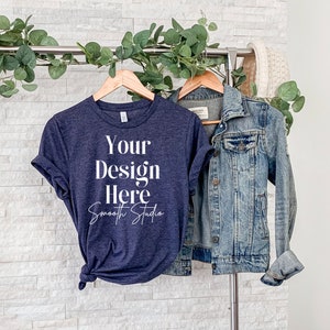 以下が含まれることがあります： 前面に「Your Design Here Smooth Studio」という文字が印刷されたネイビーブルーのTシャツ。Tシャツは木製ハンガーにかけられており、デニムジャケットがかけられています。